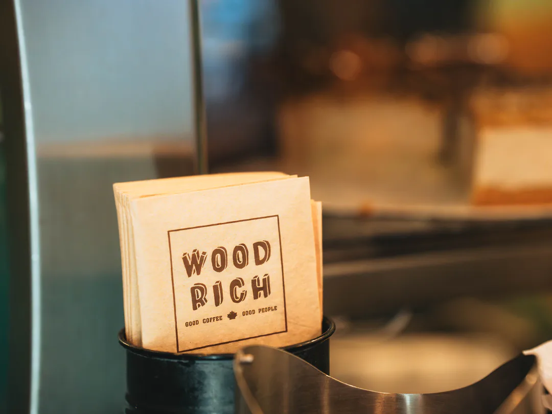 Papierservietten mit dem Aufdruck „Wood Rich – Good Coffee, Good People“ in einem schwarzen Becher