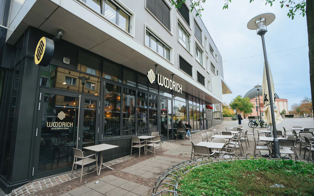 Eckfassade mit Woodrich-Schriftzug und rundem Logoschild, davor eine Terrasse mit Stühlen, hinten ein Kuppelbau